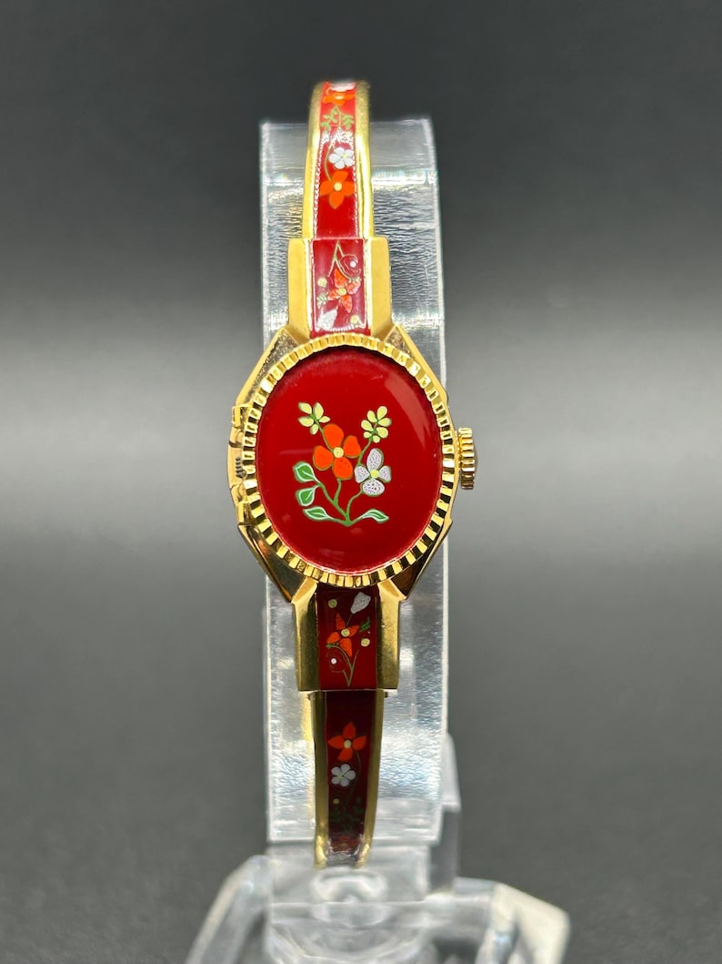 Puede incluir: Un reloj vintage dorado con esfera y correa rojas. La esfera del reloj presenta un dise&ntilde;o floral con detalles en naranja, blanco y verde. La correa tambi&eacute;n es roja con motivos florales. El reloj se exhibe sobre un soporte transparente.
