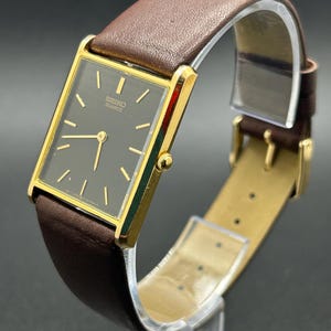 May include: A gold-toned Seiko Quartz watch with a rectangular dark gray face and gold hands. The watch has a brown leather band with a gold buckle. The watch is displayed on a clear stand against a dark background.