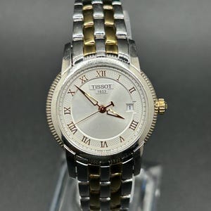 May include: A Tissot 1853 wristwatch featuring a silver-toned dial with Roman numeral hour markers, a date window at the three o'clock position, and gold-toned hands. The watch has a two-tone stainless steel link bracelet in silver and gold.