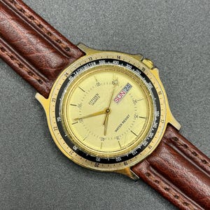 May include: A gold-toned Citizen Quartz watch with a brown leather strap. The watch face displays the day and date, with a tachymeter bezel. The watch has a water-resistant feature.