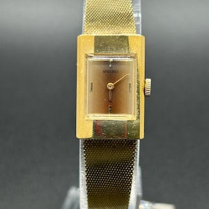 May include: A vintage gold-toned Seiko wristwatch with a rectangular face and a mesh band. The watch face is a gradient brown color with gold hands and the Seiko logo. The watch is displayed on a clear stand.