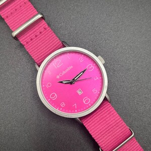 May include: A bright pink Columbia watch with a matching pink nylon strap. The watch face is pink with silver hands and numerals. The watch has a date display at the 3 o'clock position. The watch band is secured with silver-tone metal loops.