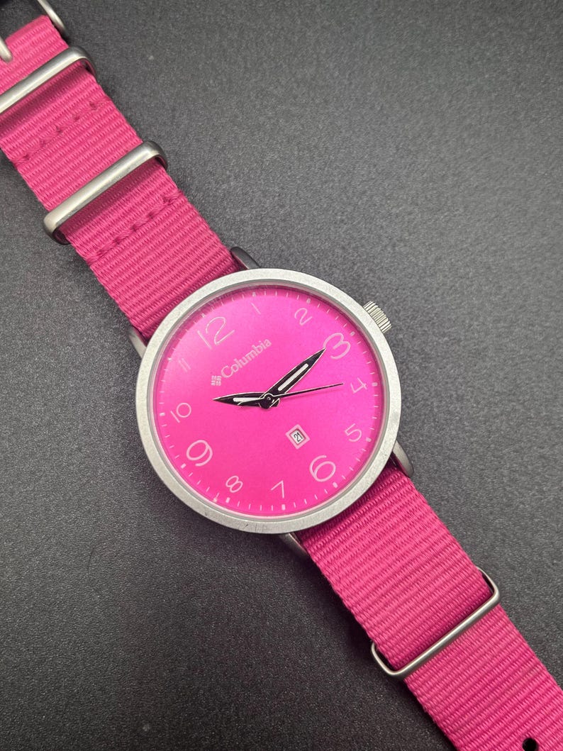 May include: A bright pink Columbia watch with a matching pink nylon strap. The watch face is pink with silver hands and numerals. The watch has a date display at the 3 o'clock position. The watch band is secured with silver-tone metal loops.