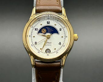 Reloj de pulsera clásico de cuarzo Timex para mujer, modelo vintage, con fase lunar, caja dorada y fecha, con correa de cuero marrón.