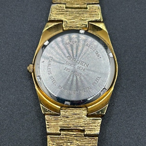 May include: A gold-tone wristwatch with a textured link band and a round case. The stainless steel case back features the text WATER RESISTANT, GRUEN SINCE 1874, 225-2035, and STAINLESS STEEL BACK BASE METAL BEZEL in a circular pattern.