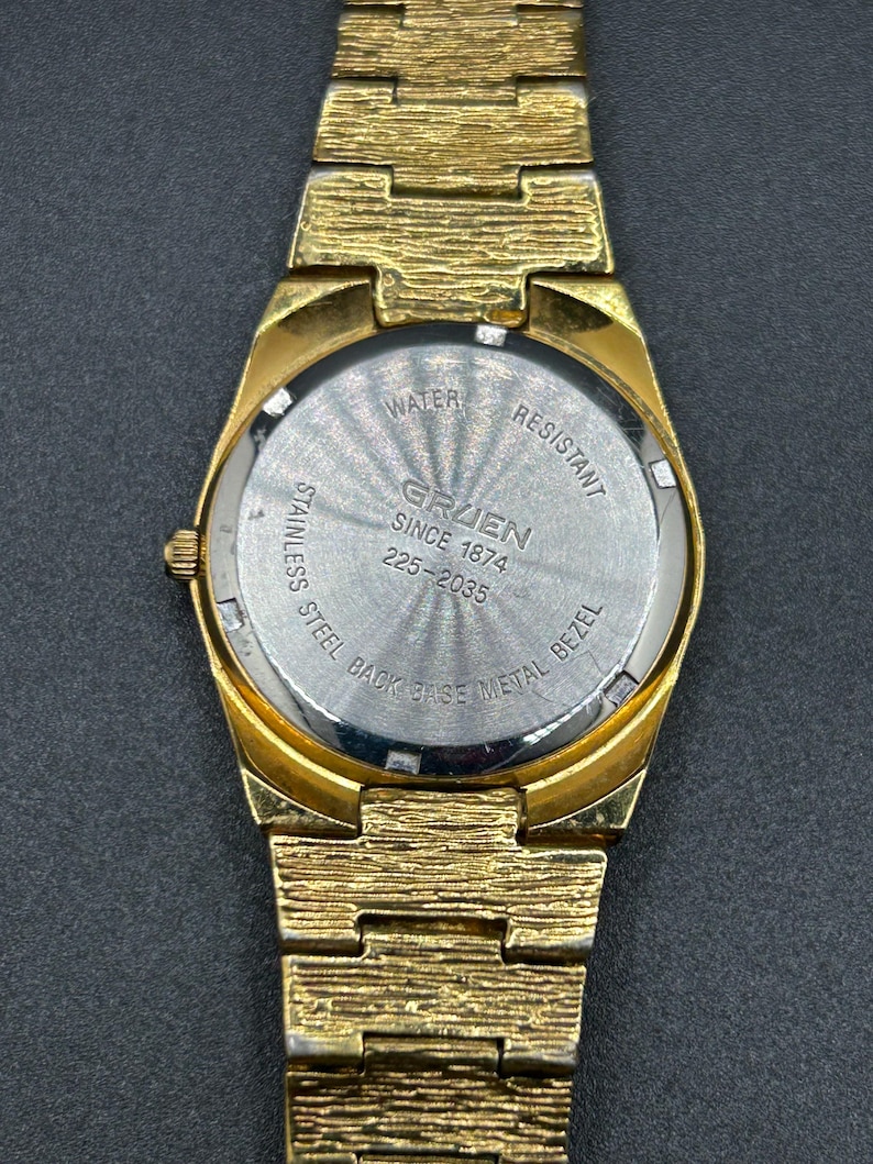 May include: A gold-tone wristwatch with a textured link band and a round case. The stainless steel case back features the text WATER RESISTANT, GRUEN SINCE 1874, 225-2035, and STAINLESS STEEL BACK BASE METAL BEZEL in a circular pattern.