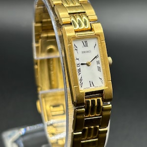 May include: A gold-toned Seiko wristwatch with a rectangular white face and black Roman numeral hour markers. The watch features a gold link band and is displayed on a clear stand. The watch face reads "SEIKO" and "JAPAN".