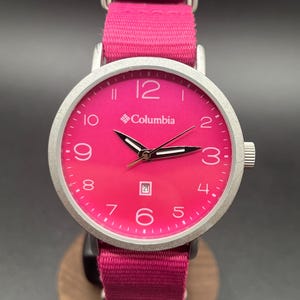 May include: A wristwatch with a bright pink face and band. The watch has a silver-colored bezel and a date display at the 6 o'clock position. The brand name "Columbia" is printed on the watch face.