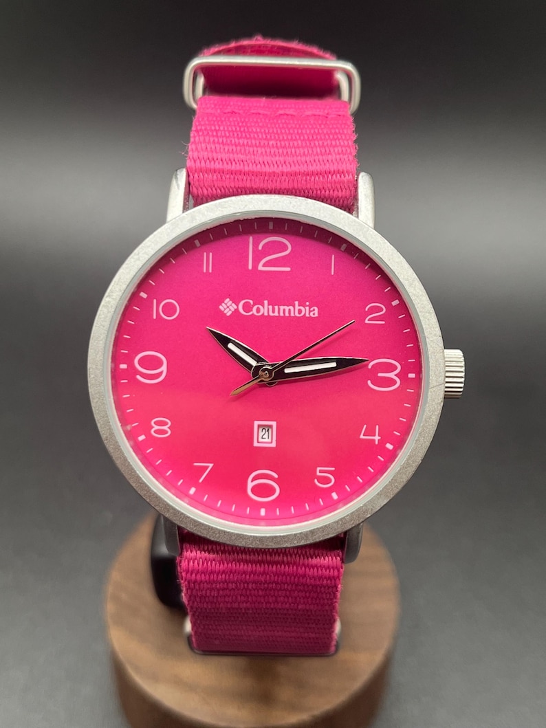 May include: A wristwatch with a bright pink face and band. The watch has a silver-colored bezel and a date display at the 6 o'clock position. The brand name "Columbia" is printed on the watch face.