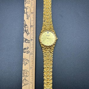 May include: A gold-tone quartz wristwatch with a textured nugget-style link band and a round face featuring the text Gruen Diamond. The watch is positioned next to a wooden ruler showing a length of approximately 7.5 inches from end to end.