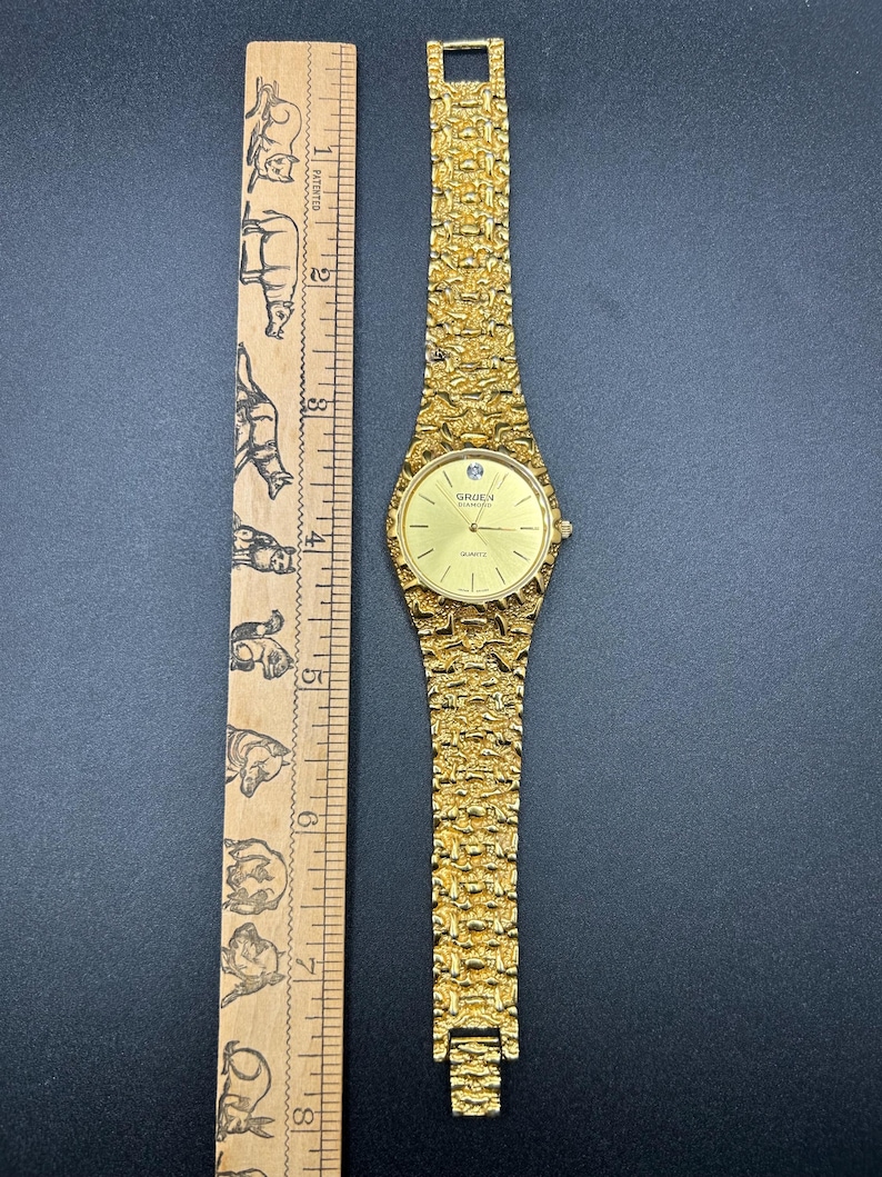 May include: A gold-tone quartz wristwatch with a textured nugget-style link band and a round face featuring the text Gruen Diamond. The watch is positioned next to a wooden ruler showing a length of approximately 7.5 inches from end to end.