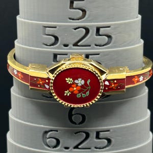 Puede incluir: Una pulsera dorada con una banda de esmalte rojo y un dise&ntilde;o circular rojo central con un motivo floral. La pulsera est&aacute; adornada con peque&ntilde;os acentos florales coloridos. La pulsera descansa sobre un medidor de anillos gris.