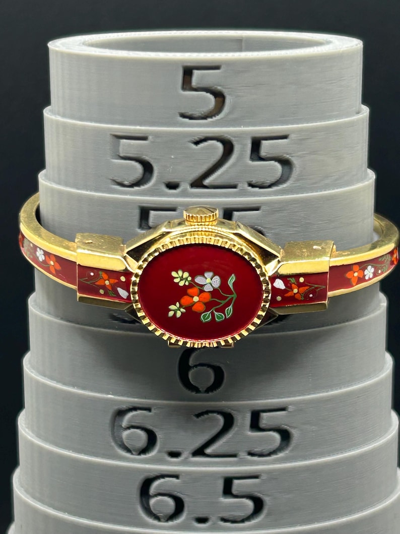 Puede incluir: Una pulsera dorada con una banda de esmalte rojo y un dise&ntilde;o circular rojo central con un motivo floral. La pulsera est&aacute; adornada con peque&ntilde;os acentos florales coloridos. La pulsera descansa sobre un medidor de anillos gris.