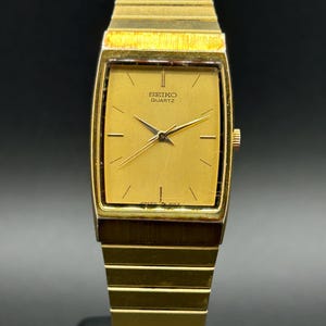 May include: A gold-toned Seiko Quartz watch with a rectangular face and a matching gold band. The watch face displays the Seiko Quartz logo and simple gold hands. The watch is a classic design.