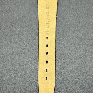 K&ouml;nnte beinhalten: Nahaufnahme eines Uhrenarmbands aus hellbraunem Kalbsleder. Der Text "GENUINE CALFSKIN" ist auf dem Armband aufgedruckt. Das Armband hat mehrere L&ouml;cher zur Anpassung und ist an einem schwarzen Zifferblatt befestigt.