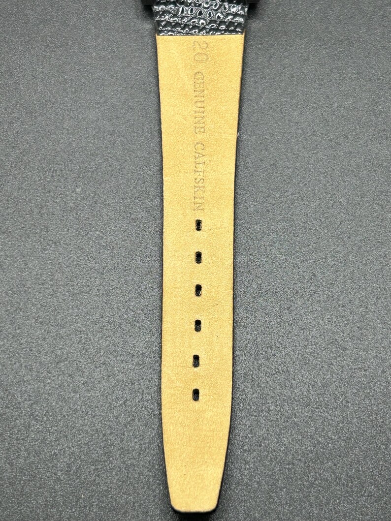 K&ouml;nnte beinhalten: Nahaufnahme eines Uhrenarmbands aus hellbraunem Kalbsleder. Der Text "GENUINE CALFSKIN" ist auf dem Armband aufgedruckt. Das Armband hat mehrere L&ouml;cher zur Anpassung und ist an einem schwarzen Zifferblatt befestigt.