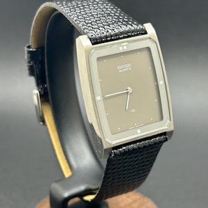 K&ouml;nnte beinhalten: Eine Vintage Ricoh Quartz-Uhr mit einem rechteckigen, silberfarbenen Geh&auml;use und einem taupefarbenen Zifferblatt. Die Uhr hat ein schwarzes, strukturiertes Lederarmband. Die Uhr wird auf einem Holzst&auml;nder pr&auml;sentiert.