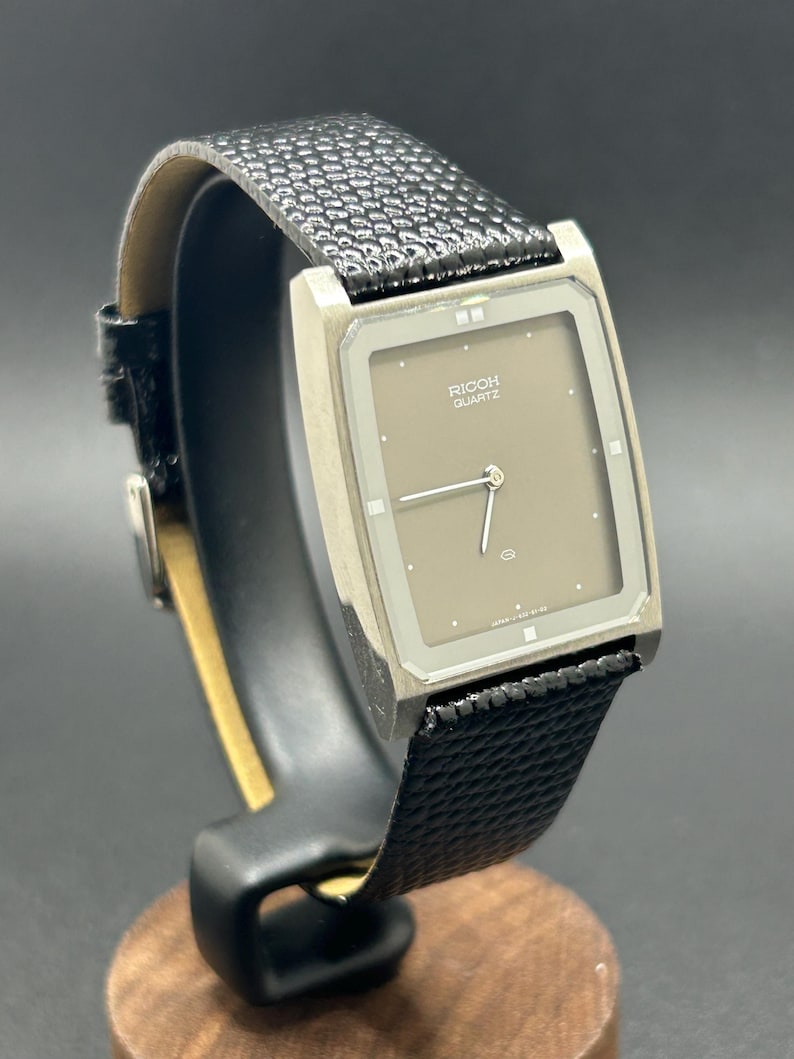 K&ouml;nnte beinhalten: Eine Vintage Ricoh Quartz-Uhr mit einem rechteckigen, silberfarbenen Geh&auml;use und einem taupefarbenen Zifferblatt. Die Uhr hat ein schwarzes, strukturiertes Lederarmband. Die Uhr wird auf einem Holzst&auml;nder pr&auml;sentiert.