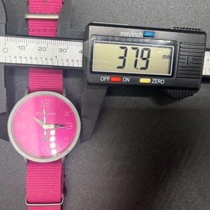 May include: A pink Columbia watch with a matching pink fabric band is measured with a digital caliper. The watch face is pink with silver hands and hour markers. The caliper displays a measurement of 37.9 mm.