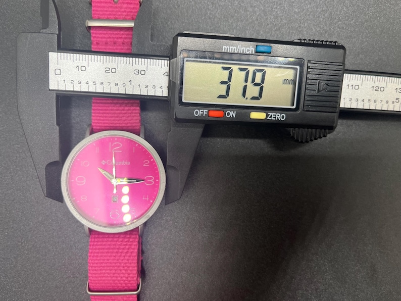 May include: A pink Columbia watch with a matching pink fabric band is measured with a digital caliper. The watch face is pink with silver hands and hour markers. The caliper displays a measurement of 37.9 mm.