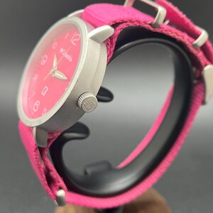 May include: A silver-toned Columbia watch with a bright pink face and matching fabric band. The watch features a round face with white numerals and hands. The band is secured with silver-toned metal hardware.