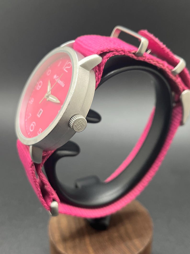 May include: A silver-toned Columbia watch with a bright pink face and matching fabric band. The watch features a round face with white numerals and hands. The band is secured with silver-toned metal hardware.
