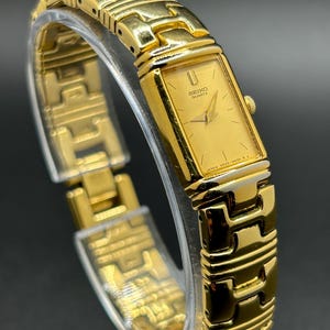 May include: A gold-toned Seiko Quartz watch with a rectangular face and a matching link bracelet. The watch face displays the Seiko logo and the word "Quartz". The watch is displayed on a clear stand.