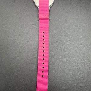 May include: A wristwatch with a silver-colored case and a bright pink nylon strap. The strap has multiple holes for adjustment. The watch is set against a dark background.