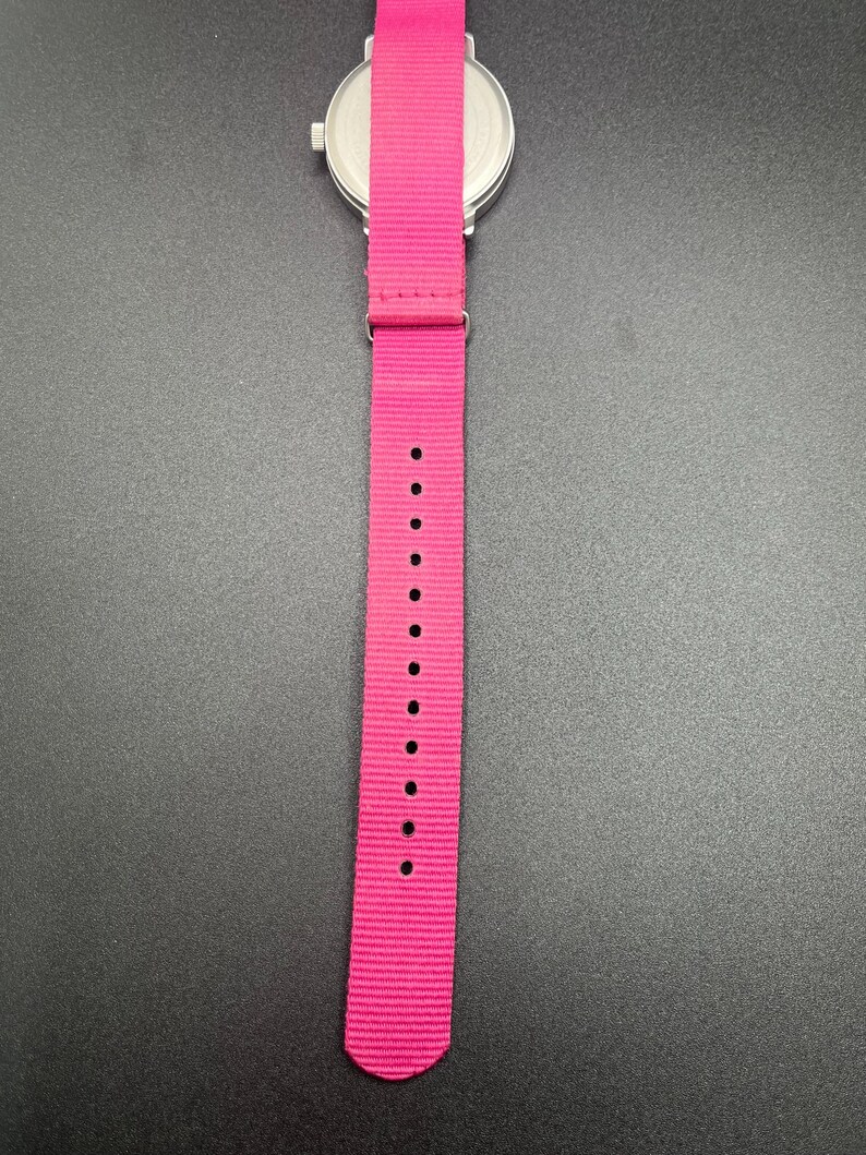 May include: A wristwatch with a silver-colored case and a bright pink nylon strap. The strap has multiple holes for adjustment. The watch is set against a dark background.