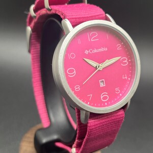 May include: A pink Columbia watch with a silver-tone case and a matching pink fabric band. The watch face features white numerals and hands, with the brand name printed below the 12. The band is secured with silver-tone metal loops.