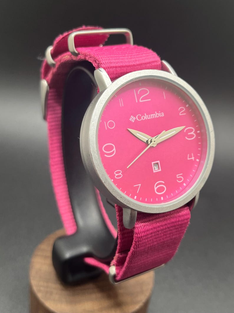 May include: A pink Columbia watch with a silver-tone case and a matching pink fabric band. The watch face features white numerals and hands, with the brand name printed below the 12. The band is secured with silver-tone metal loops.