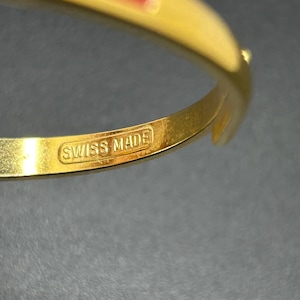 Puede incluir: Una pulsera dorada con una franja roja. La pulsera tiene un cierre y el texto "SWISS MADE" es visible. La joya est&aacute; sobre una superficie oscura.