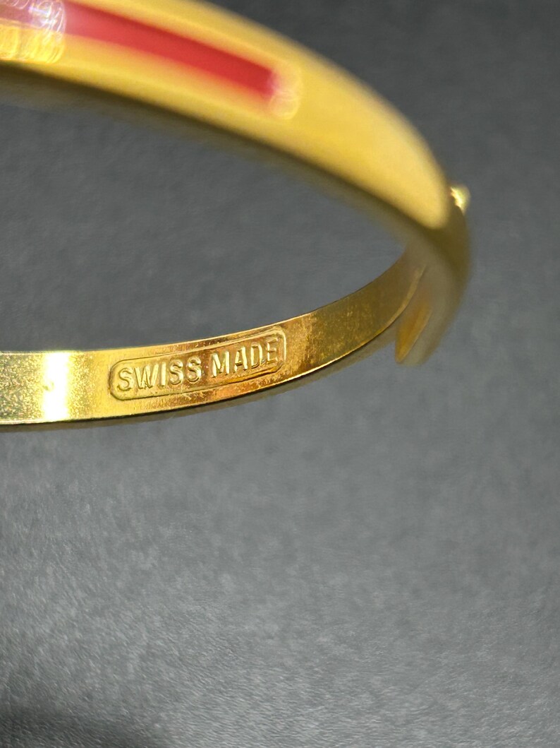 Puede incluir: Una pulsera dorada con una franja roja. La pulsera tiene un cierre y el texto "SWISS MADE" es visible. La joya est&aacute; sobre una superficie oscura.