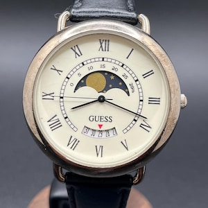 May include: A silver Guess watch with a cream face and black leather strap. The watch face displays Roman numerals, a moon phase indicator, and the Guess logo. The watch is presented on a wooden stand.