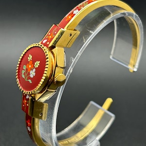 Puede incluir: Un reloj de pulsera dorado con esfera y correa rojas, adornado con un dise&ntilde;o floral. La esfera del reloj es ovalada, con un fondo rojo y flores blancas, naranjas y verdes. La correa tambi&eacute;n es roja con un estampado floral.