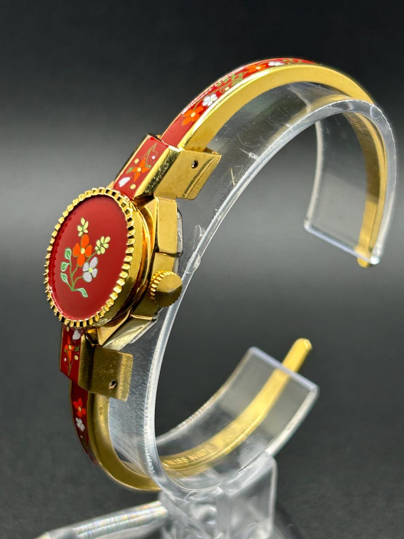 Puede incluir: Un reloj de pulsera dorado con esfera y correa rojas, adornado con un dise&ntilde;o floral. La esfera del reloj es ovalada, con un fondo rojo y flores blancas, naranjas y verdes. La correa tambi&eacute;n es roja con un estampado floral.