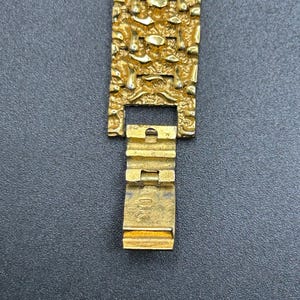 May include: A gold-colored metal watch band featuring a textured, nugget-like surface pattern. The band includes a hinged clasp mechanism with the letters DK clearly engraved on the interior surface of the folding metal closure component.