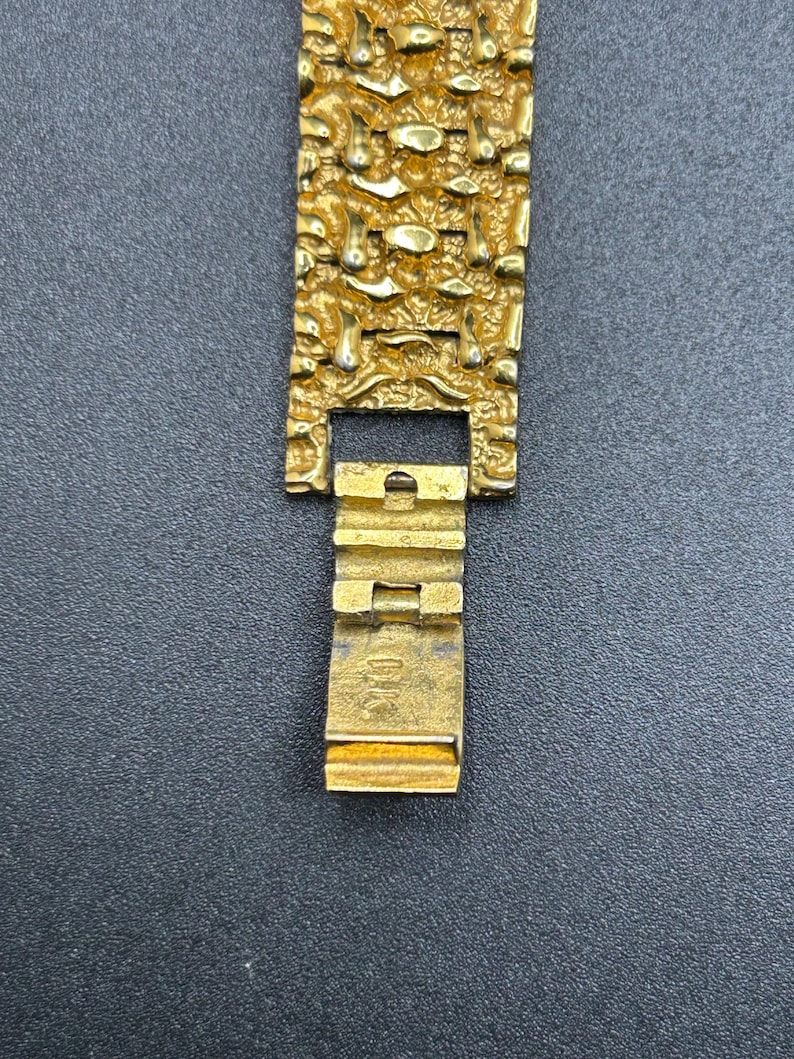May include: A gold-colored metal watch band featuring a textured, nugget-like surface pattern. The band includes a hinged clasp mechanism with the letters DK clearly engraved on the interior surface of the folding metal closure component.