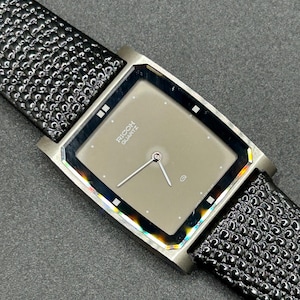 K&ouml;nnte beinhalten: Eine rechteckige Armbanduhr mit grauem Zifferblatt und silbernen Zeigern. Auf dem Zifferblatt steht "RICOH QUARTZ". Die Uhr hat ein schwarzes, strukturiertes Lederarmband.