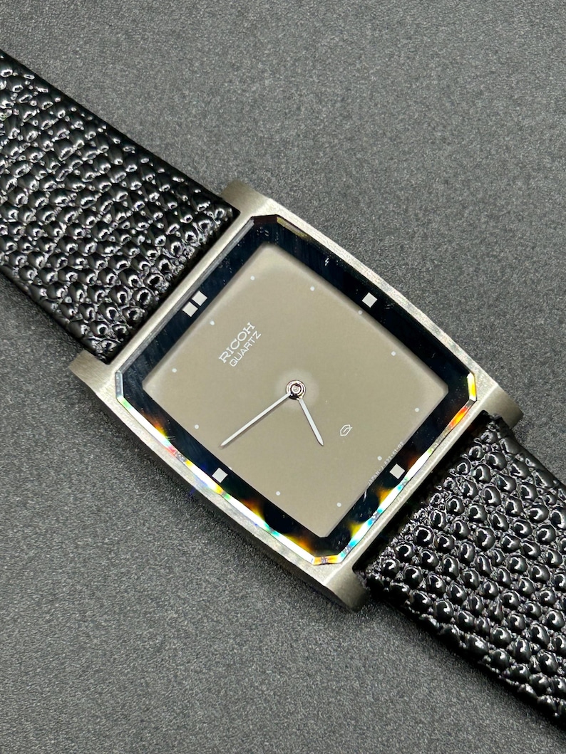 K&ouml;nnte beinhalten: Eine rechteckige Armbanduhr mit grauem Zifferblatt und silbernen Zeigern. Auf dem Zifferblatt steht "RICOH QUARTZ". Die Uhr hat ein schwarzes, strukturiertes Lederarmband.