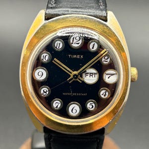 May include: Vintage Timex watch with a black face, gold-tone bezel, and black leather band. The watch face displays white numerals, gold hands, and the day of the week. The watch is water resistant.