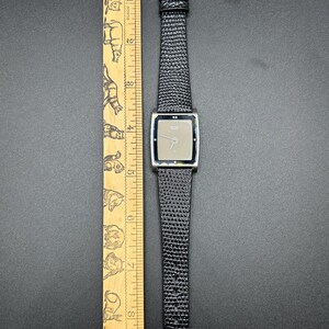 K&ouml;nnte beinhalten: Eine silberfarbene Armbanduhr mit rechteckigem Zifferblatt und einem schwarzen, strukturierten Lederarmband. Das Zifferblatt der Uhr ist hellgrau. Ein Holzlineal mit Tierillustrationen liegt neben der Uhr.