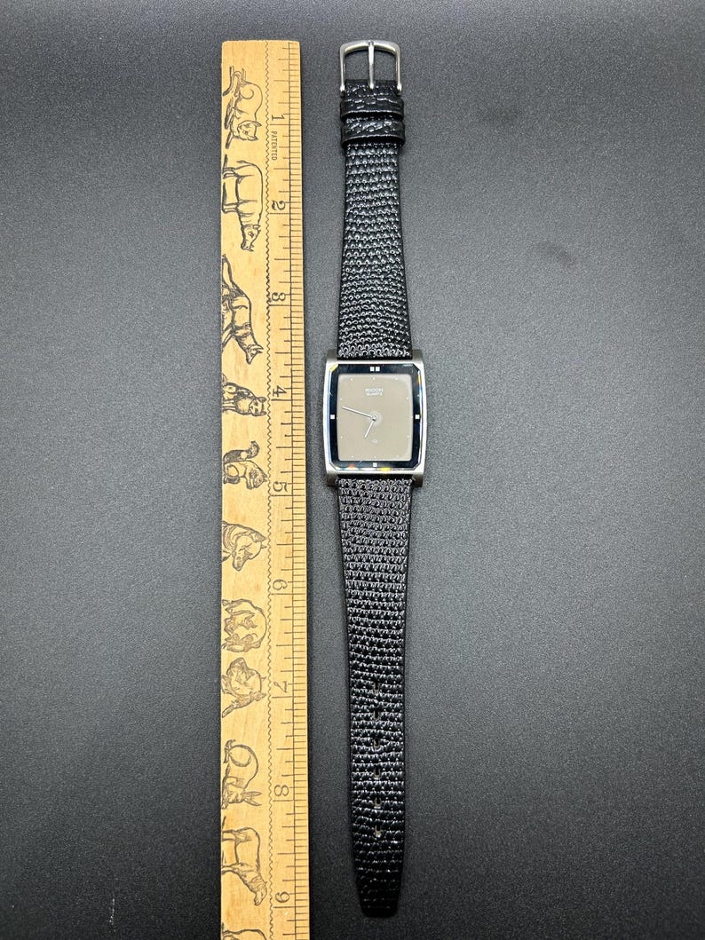 K&ouml;nnte beinhalten: Eine silberfarbene Armbanduhr mit rechteckigem Zifferblatt und einem schwarzen, strukturierten Lederarmband. Das Zifferblatt der Uhr ist hellgrau. Ein Holzlineal mit Tierillustrationen liegt neben der Uhr.