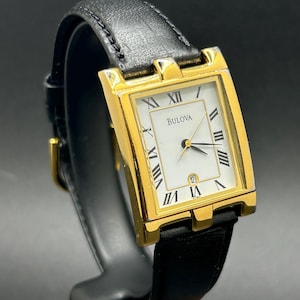May include: A rectangular gold-tone Bulova wristwatch featuring a white dial with black Roman numeral hour markers, three thin hands, and a date window at the six o'clock position. The watch is attached to a black leather strap with a gold buckle.