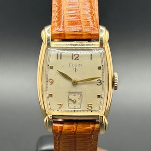 May include: A vintage Elgin wristwatch with a gold-tone rectangular case and a cream-coloured face. The watch features gold hands and hour markers, a small seconds subdial, and a brown leather strap. The watch face displays the brand name "Elgin".