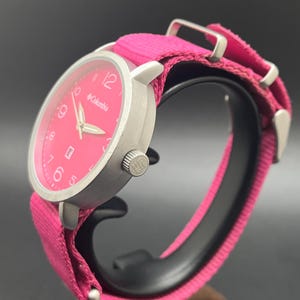May include: A silver-toned Columbia watch with a bright pink face and matching fabric band. The watch features a round face with white numerals and hands, and the Columbia logo. The band is secured with silver-toned metal hardware.