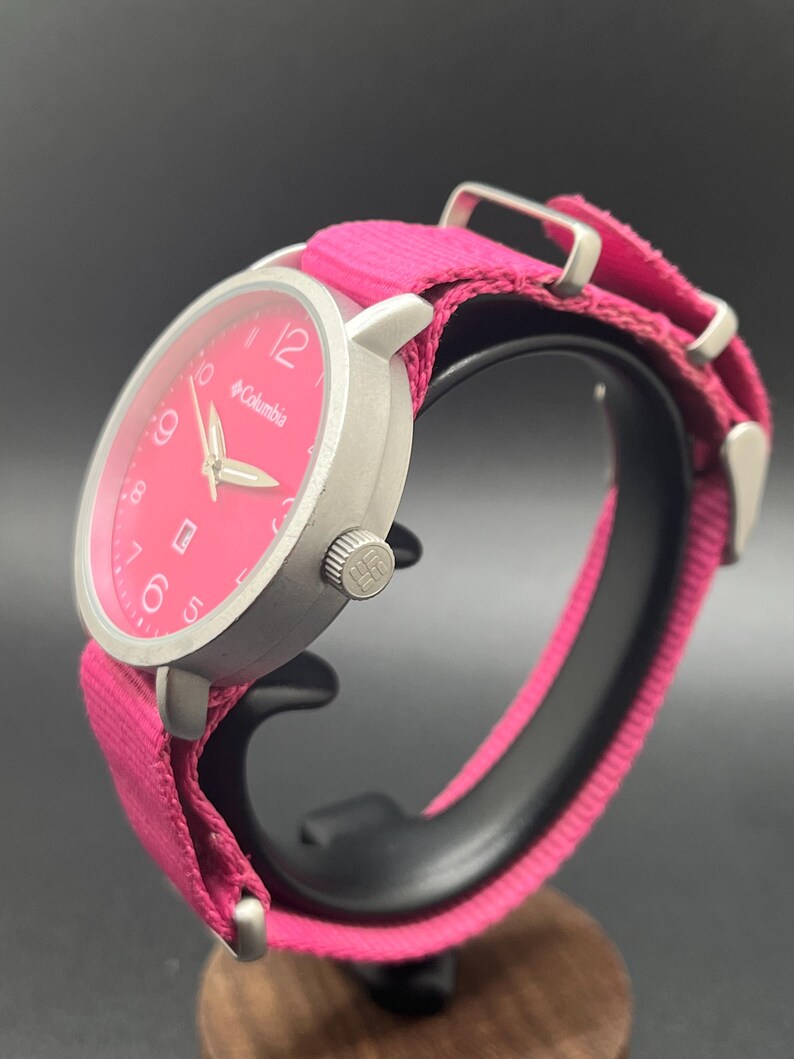 May include: A silver-toned Columbia watch with a bright pink face and matching fabric band. The watch features a round face with white numerals and hands, and the Columbia logo. The band is secured with silver-toned metal hardware.