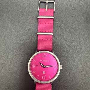 May include: A Columbia watch with a bright pink face and matching pink nylon band. The watch has a silver-colored case and buckle. The watch face displays the Columbia logo, numerals, and hands.