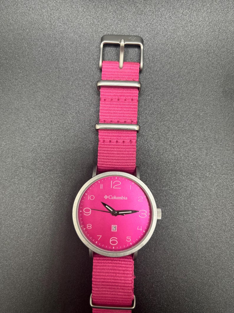 May include: A Columbia watch with a bright pink face and matching pink nylon band. The watch has a silver-colored case and buckle. The watch face displays the Columbia logo, numerals, and hands.