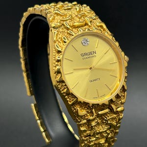 May include: A gold-tone Gruen Diamond quartz watch featuring a textured, nugget-style link bracelet and a round gold dial. The watch face displays the text Gruen Diamond and Quartz, with a single diamond marker at the twelve o'clock position.